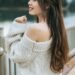 Woman wearing an oversized off-white knit sweater, standing outdoors by a lake railing, smiling softly with her hair cascading down, evoking cozy, casual, and timeless fall wardrobe style.