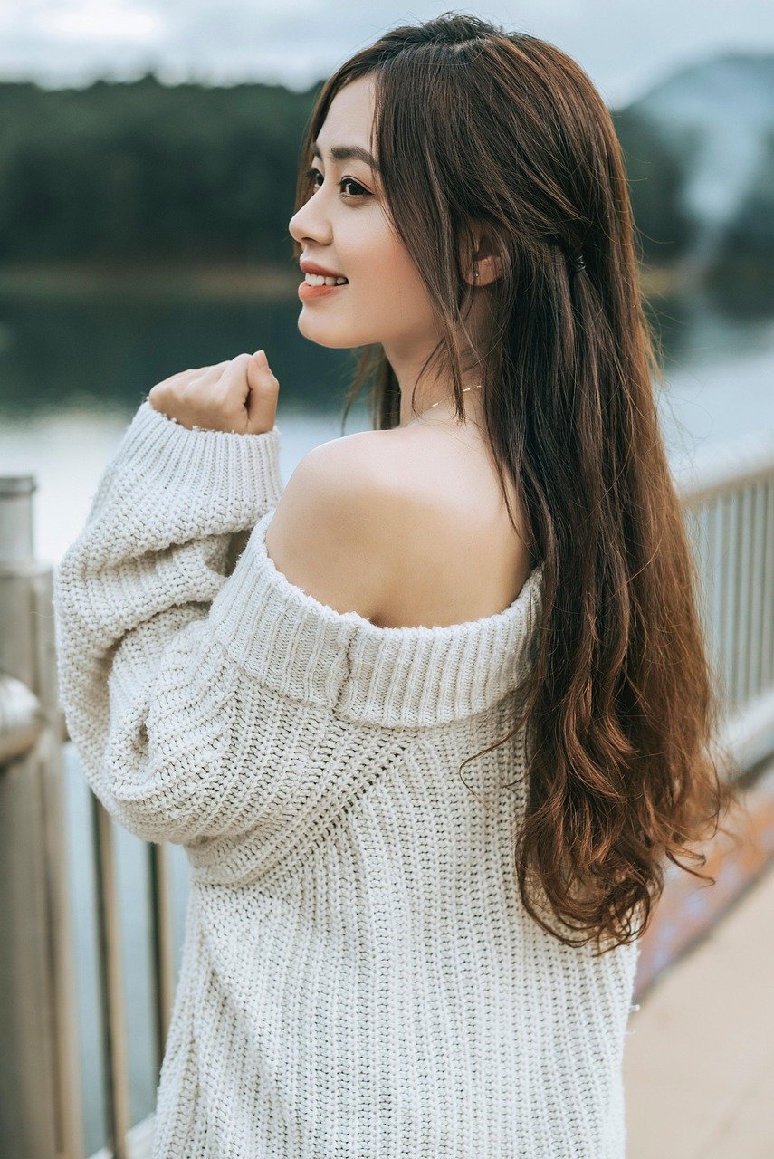Woman wearing an oversized off-white knit sweater, standing outdoors by a lake railing, smiling softly with her hair cascading down, evoking cozy, casual, and timeless fall wardrobe style.