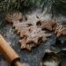 Close-up of freshly baked Christmas cookies in star and tree shapes, dusted with powdered sugar. A wooden rolling pin and star-shaped cookie cutters are nearby, with pine branches in the background, creating a festive holiday baking scene.