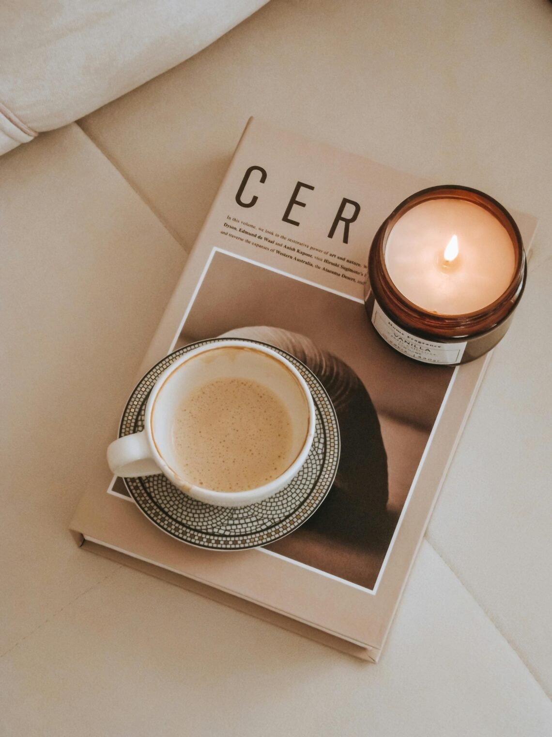 Overhead flat lay of a cozy scene with a cup of coffee on a patterned saucer resting on a neutral-toned magazine, accompanied by a lit brown jar candle, highlighting calm, comfortable home essentials for a Cyber Monday Amazon feature.