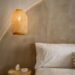 Minimalist bedroom corner with a white bed, soft bedding, and a round wooden side table. A woven pendant light hangs above, casting a warm glow against the neutral textured wall, creating a serene and cozy evening atmosphere.