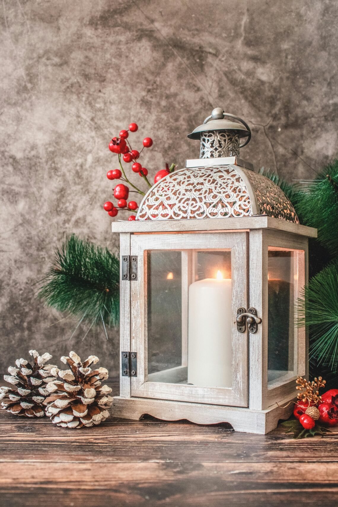 White metal Christmas lantern with intricate filigree top, lit candle inside, surrounded by pine branches, red berries, and pinecones on a wooden surface. The festive setup evokes cozy holiday ambiance and rustic seasonal decor.