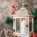 White metal Christmas lantern with intricate filigree top, lit candle inside, surrounded by pine branches, red berries, and pinecones on a wooden surface. The festive setup evokes cozy holiday ambiance and rustic seasonal decor.