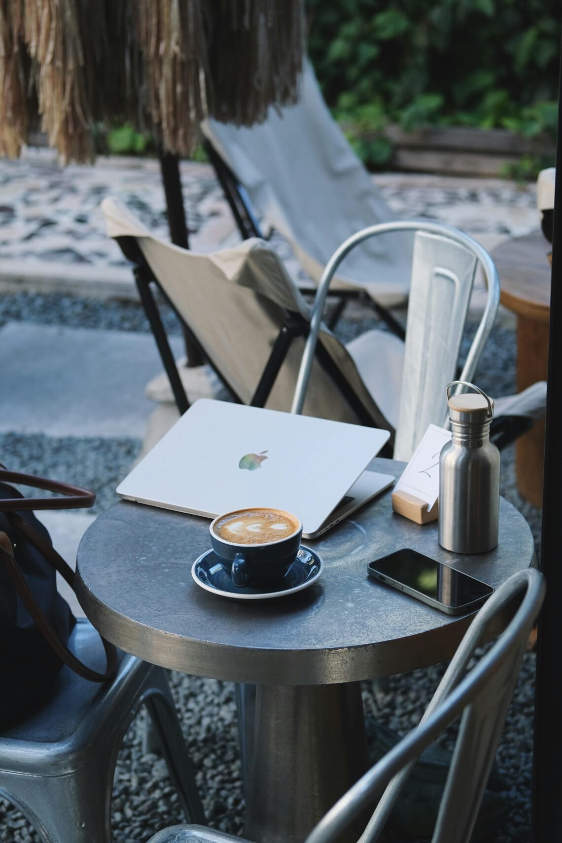 Outdoor café scene with a round metal table holding a laptop, stainless steel water bottle, smartphone, and a cup of coffee with latte art. Neutral-toned chairs and gravel patio in the background evoke a relaxed, cozy workspace or coffee break setting.