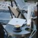 Outdoor café scene with a round metal table holding a laptop, stainless steel water bottle, smartphone, and a cup of coffee with latte art. Neutral-toned chairs and gravel patio in the background evoke a relaxed, cozy workspace or coffee break setting.