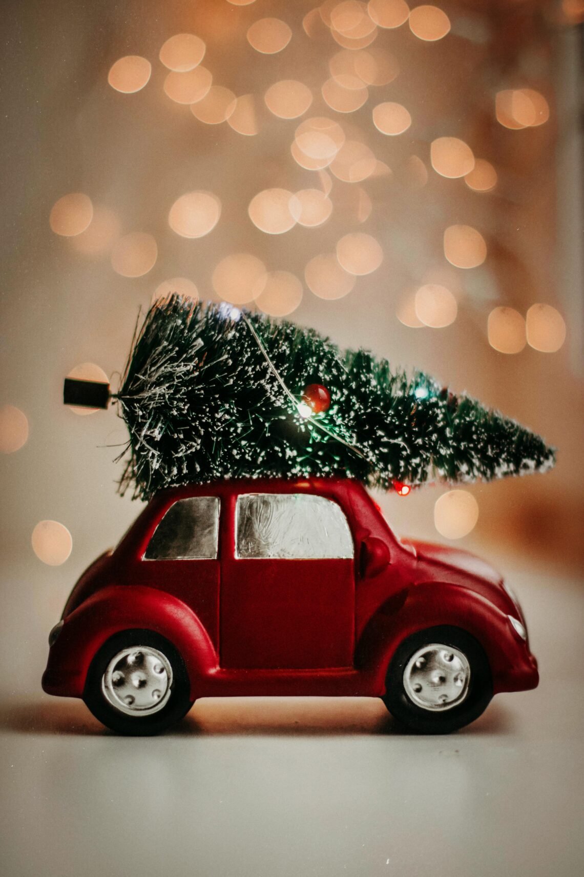 Red toy car carrying a miniature Christmas tree with blurred warm bokeh lights in the background, evoking cozy holiday gift ideas and festive Amazon wishlist inspiration