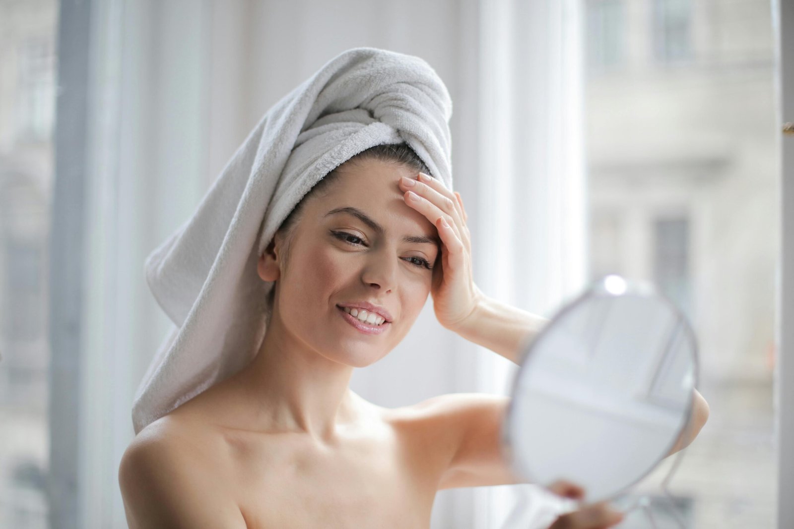 A young woman with a white towel wrapped around her hair gently touches her forehead while looking into a handheld mirror, illustrating a morning skincare routine emphasizing sunscreen application and self-care.