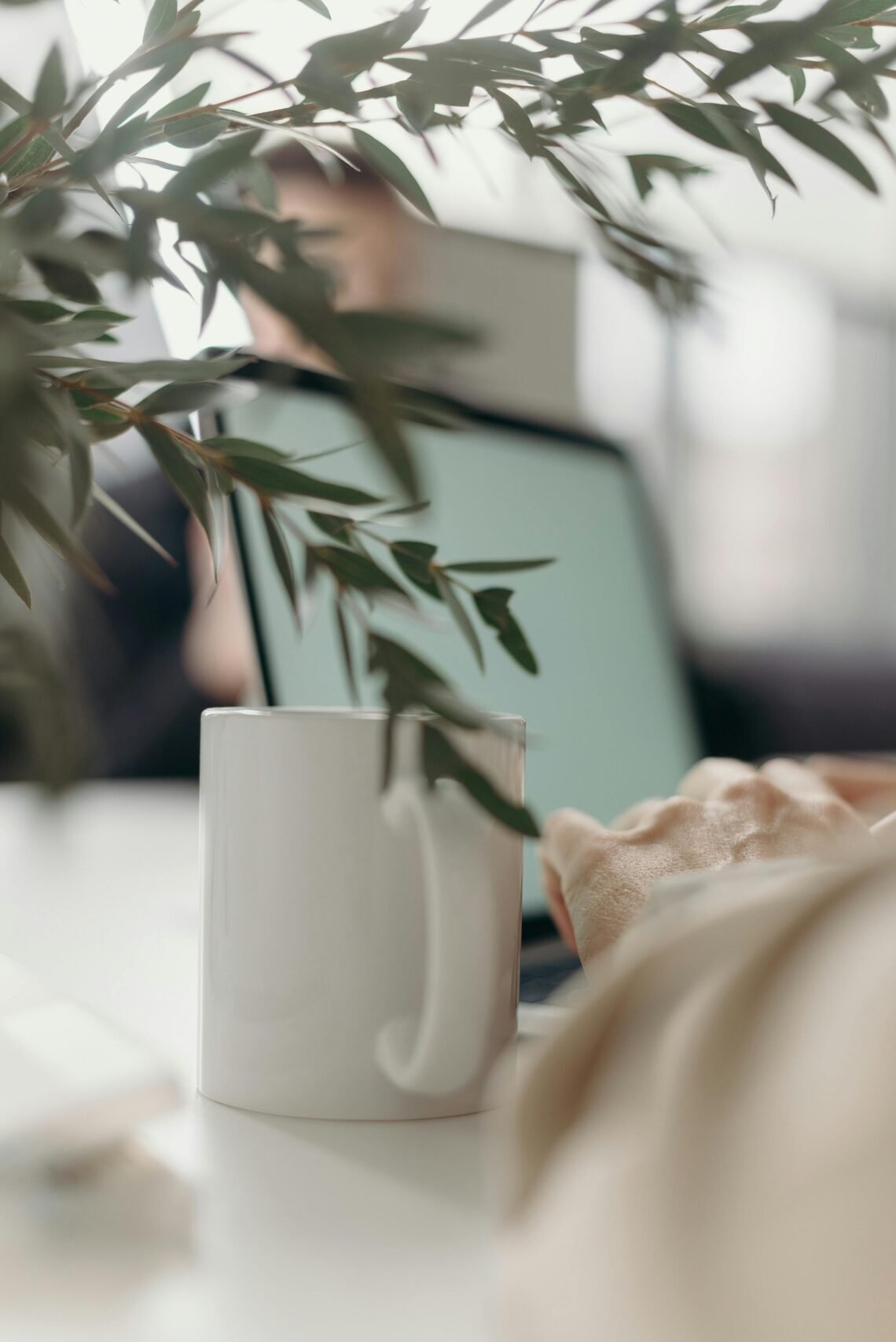 Close-up of a white coffee mug on a desk in a calm, cozy workspace, with blurred laptop and greenery in the background, evoking focus and relaxed productivity.