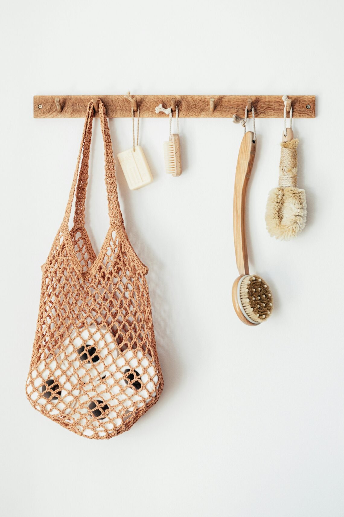 A wooden wall hook holds a hanging macramé bag filled with bath sponges, alongside a soap bar, small brush, long-handled scrub brush, and natural bristle brush, illustrating a cozy, minimalist bathroom setup with functional, everyday essentials.