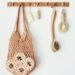 A wooden wall hook holds a hanging macramé bag filled with bath sponges, alongside a soap bar, small brush, long-handled scrub brush, and natural bristle brush, illustrating a cozy, minimalist bathroom setup with functional, everyday essentials.
