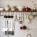 Organized kitchen with white subway tile backsplash and floating shelves, featuring patterned bowls, a colander, red cookware, glass bottles, and hanging utensils. Below, a white sink, small plants in red mugs, a wicker basket, and assorted mini pumpkins create a bright, cozy, and functional countertop setup.