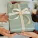 Two hands exchanging a neatly wrapped gift with sage green paper and a cream ribbon bow, accented with delicate white flowers. The background shows a cozy, blurred indoor setting, evoking a warm, thoughtful holiday or last-minute Christmas gift-giving moment.