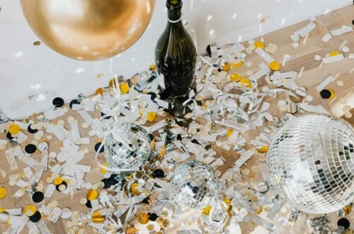 Post-party New Year’s Eve scene with gold balloon, champagne bottle, silver disco balls, and scattered confetti on a wooden floor, evoking festive celebration and sparkling holiday vibes.