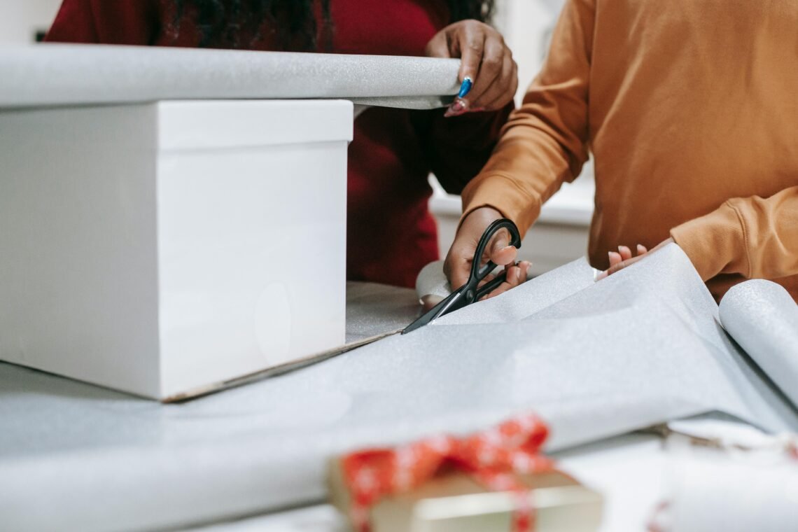 Two people wrapping a Christmas gift: one holds a white box while the other cuts silver wrapping paper with scissors. The scene highlights holiday preparation, cozy home activities, and festive gift-giving.