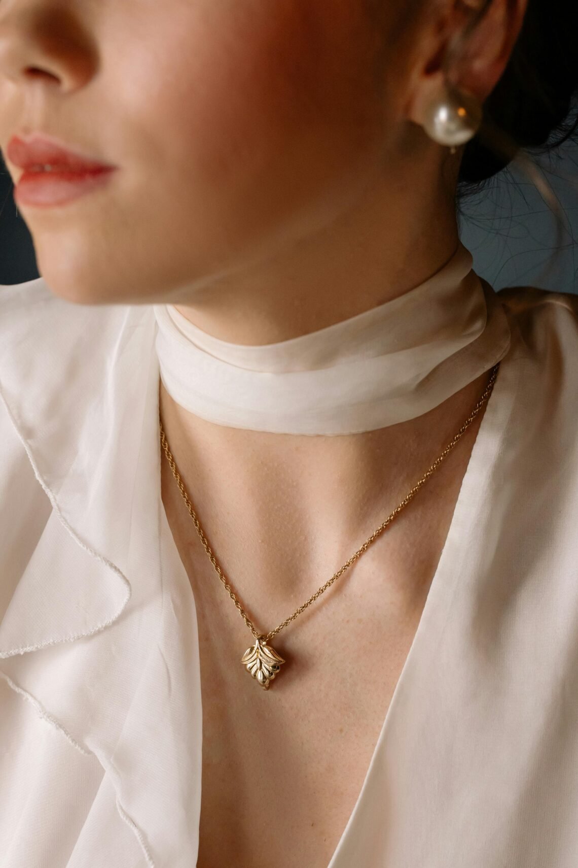 Close-up of a woman wearing a delicate gold pendant necklace and a pearl stud earring, styled with a sheer white blouse and scarf, highlighting elegant holiday jewelry for a luxury Christmas gift feature.