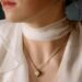 Close-up of a woman wearing a delicate gold pendant necklace and a pearl stud earring, styled with a sheer white blouse and scarf, highlighting elegant holiday jewelry for a luxury Christmas gift feature.