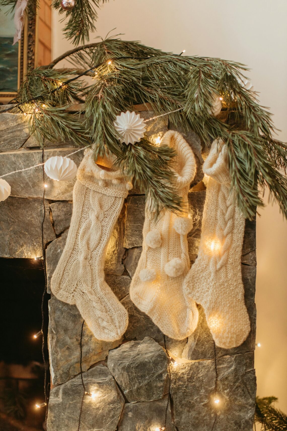 Three cream-colored knit Christmas stockings hanging from a stone fireplace mantel, adorned with greenery and warm string lights, creating a cozy and festive winter ambiance.