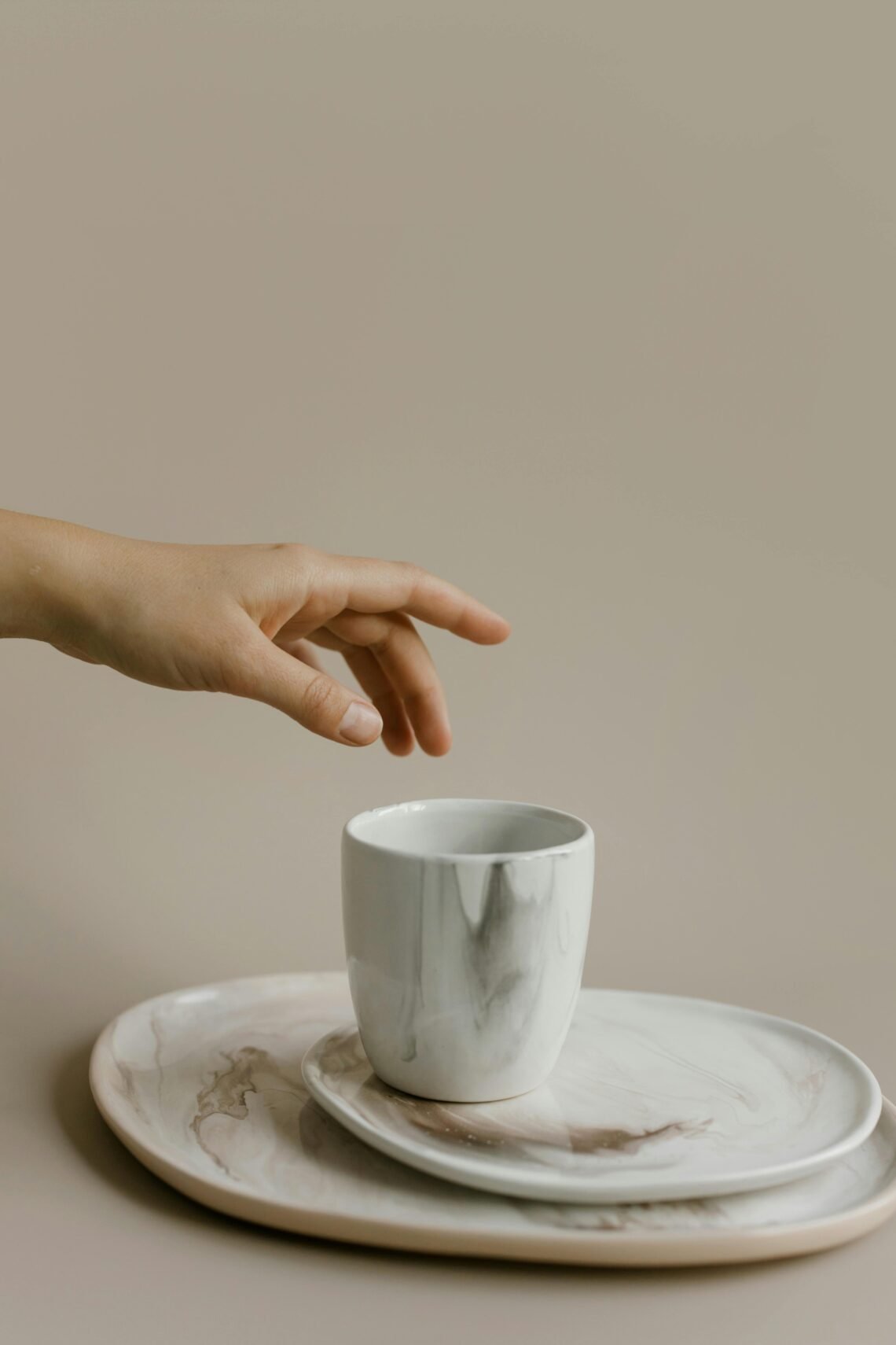 A hand reaching toward a white ceramic marble-patterned mug placed on a matching set of two round plates, evoking a calm and minimalist cozy lifestyle aesthetic.
