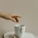 A hand reaching toward a white ceramic marble-patterned mug placed on a matching set of two round plates, evoking a calm and minimalist cozy lifestyle aesthetic.