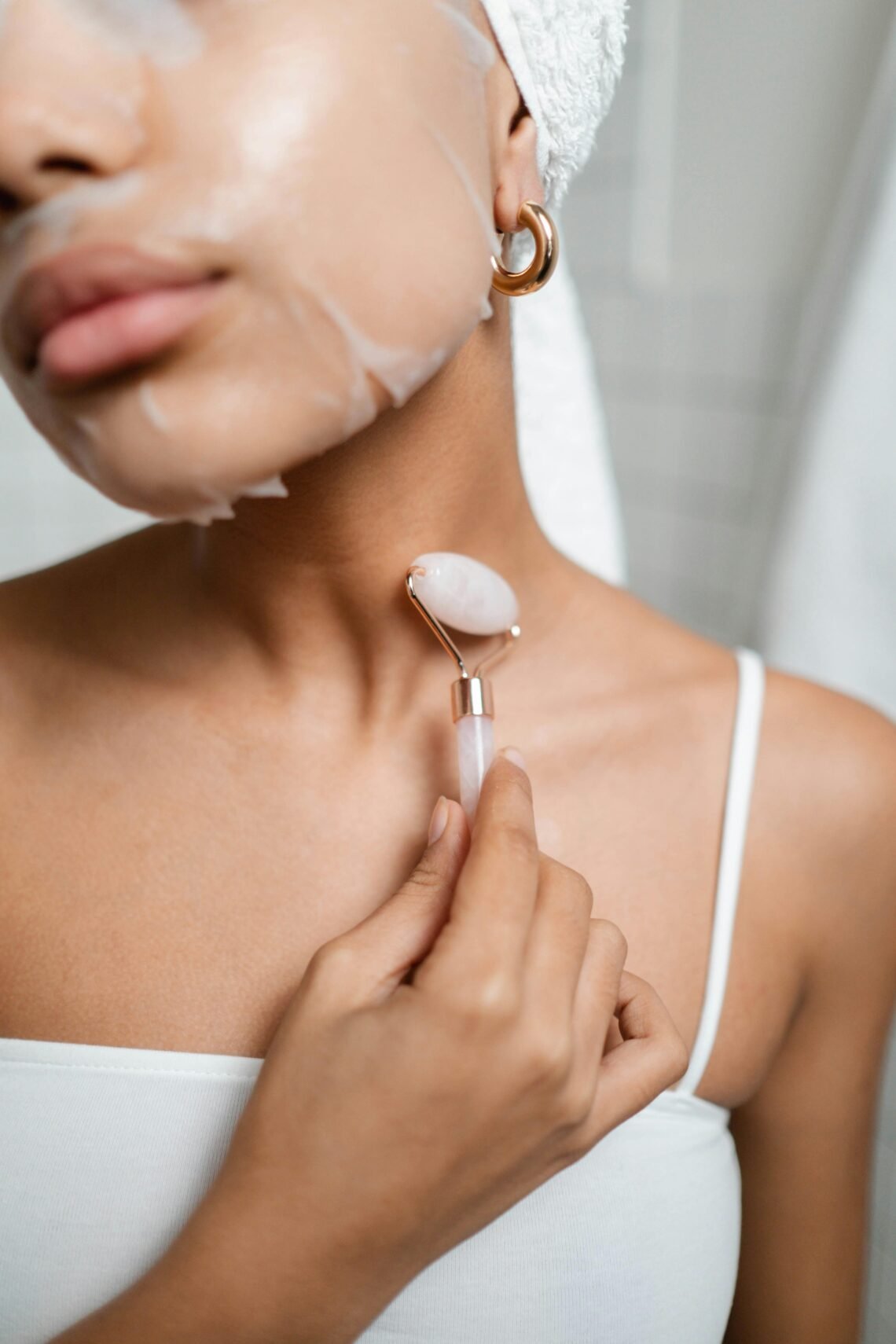 Close-up of a person using a rose quartz facial roller on their neck while wearing a white tank top and towel turban, with a hydrating sheet mask on the face, evoking calm, at-home skincare self-care routine.