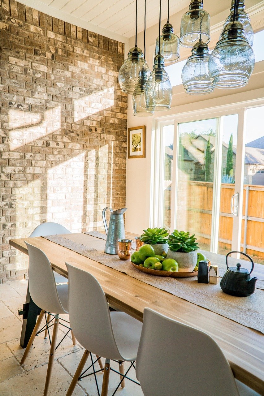 Bright and cozy breakfast nook with a long wooden dining table, white modern chairs, brick accent wall, natural light streaming through sliding glass doors, farmhouse-style hanging glass pendant lights, green apples and succulents as centerpiece, styled for a mindful meal corner and peaceful morning tea.