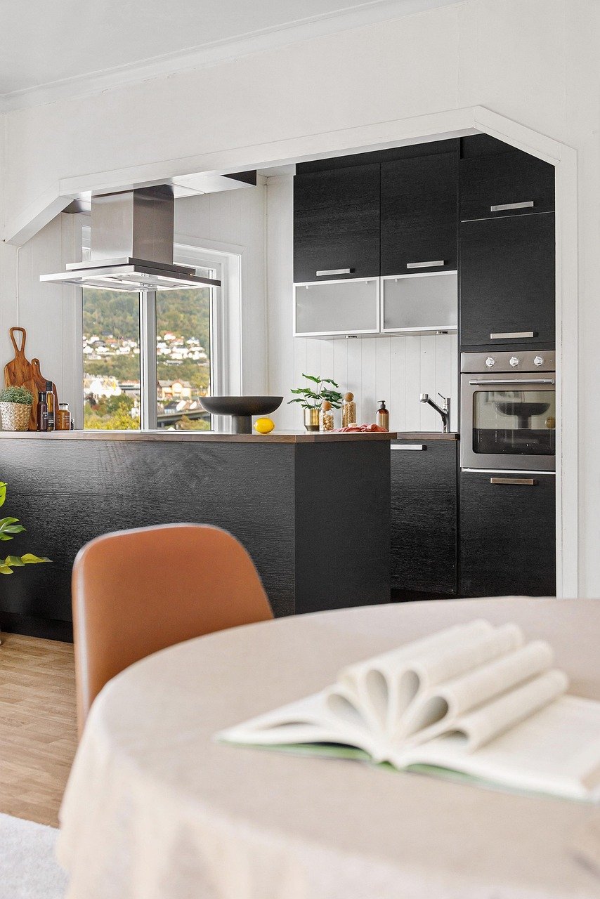 Modern small apartment kitchen with black cabinets, stainless steel appliances, large window with natural light, sleek wood countertop with plants and cutting boards, minimalist décor, and a cozy dining corner in the foreground with a blurred book, designed for maximizing space and creating a calm, serene living environment.