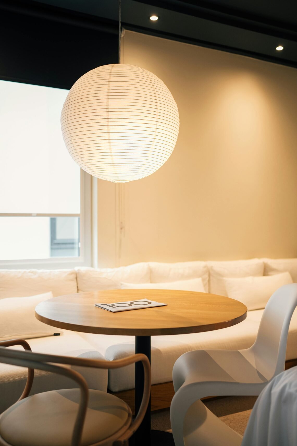Minimalist small apartment dining nook with round wood table, modern white and tan chairs, soft ambient light from hanging paper lantern pendant, neutral-colored walls, and natural light from window creating a warm and cozy atmosphere perfect for relaxing, reading, or enjoying tea.