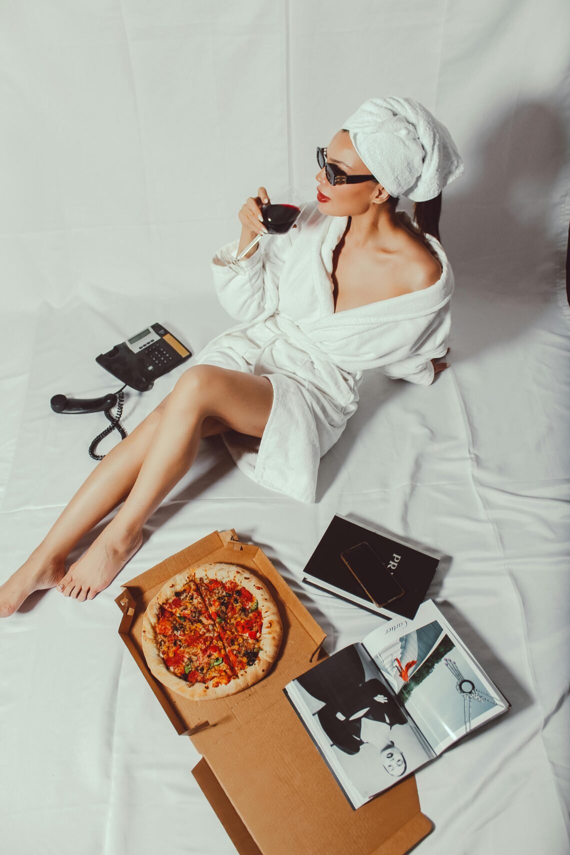 Woman in bathrobe with towel sipping wine and enjoying pizza while reading in a cozy setting.