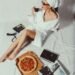 Woman in bathrobe with towel sipping wine and enjoying pizza while reading in a cozy setting.