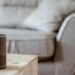 A modern smart speaker sits on a wooden table in a cozy, minimalist living room setting.