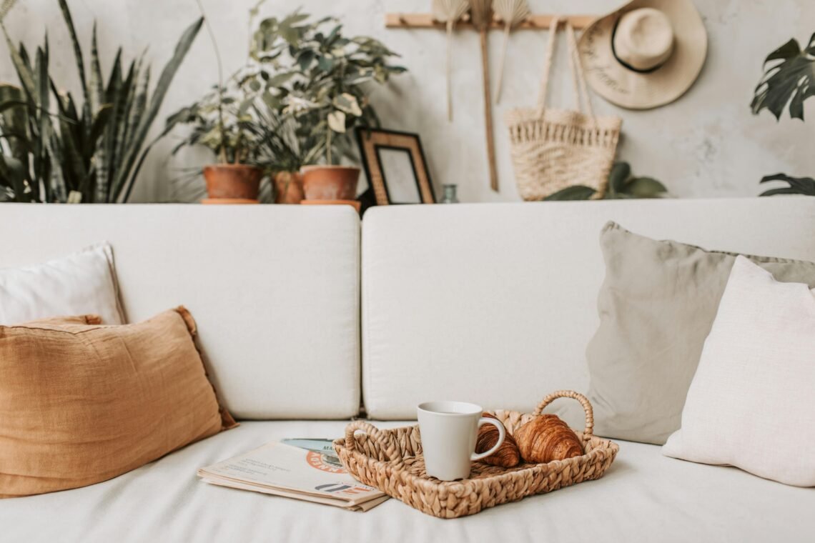 Cozy living room accent pieces styled on a white sofa with neutral pillows, a woven tray, coffee mug, greenery, and warm natural decor for a soft, inviting home look